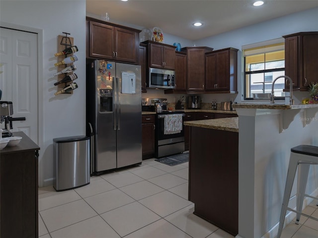 kitchen with dark brown cabinetry, stone countertops, a breakfast bar area, light tile patterned floors, and appliances with stainless steel finishes