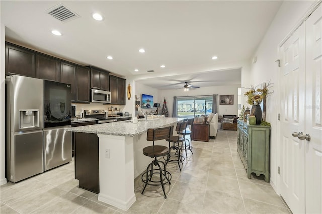 kitchen with light stone countertops, an island with sink, dark brown cabinets, a kitchen bar, and stainless steel appliances