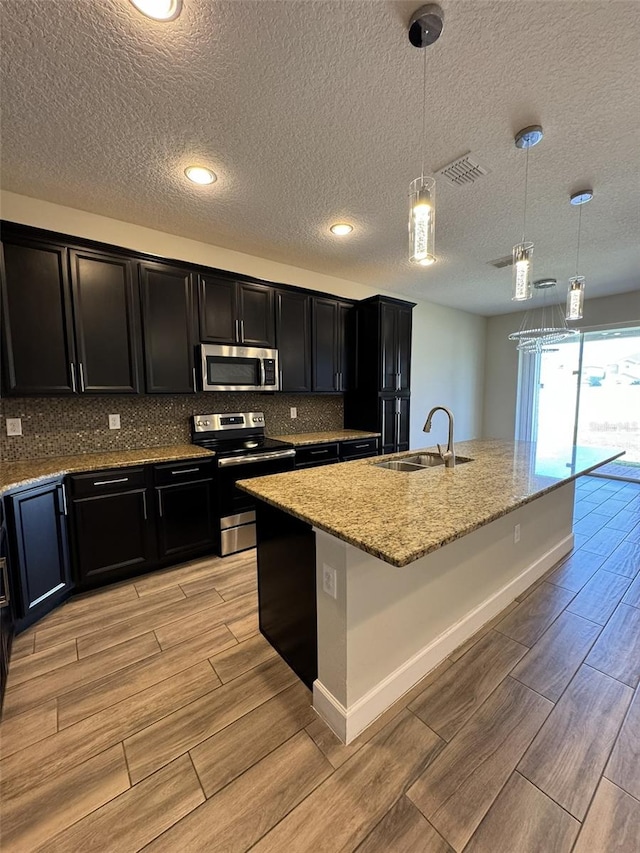 kitchen with sink, light hardwood / wood-style flooring, an island with sink, decorative light fixtures, and appliances with stainless steel finishes