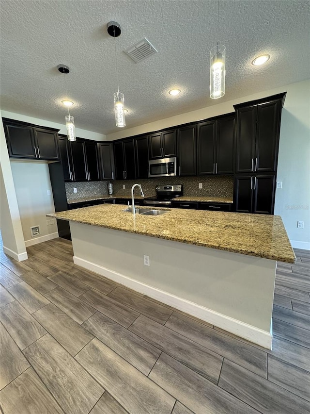 kitchen featuring appliances with stainless steel finishes, a textured ceiling, decorative light fixtures, and a center island with sink