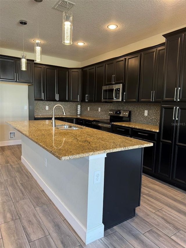 kitchen with backsplash, stainless steel appliances, decorative light fixtures, a center island with sink, and light hardwood / wood-style floors