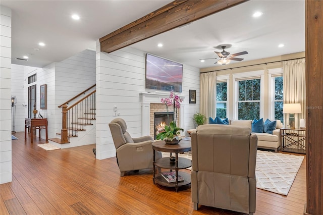 living room with ceiling fan, a brick fireplace, beamed ceiling, hardwood / wood-style floors, and wooden walls