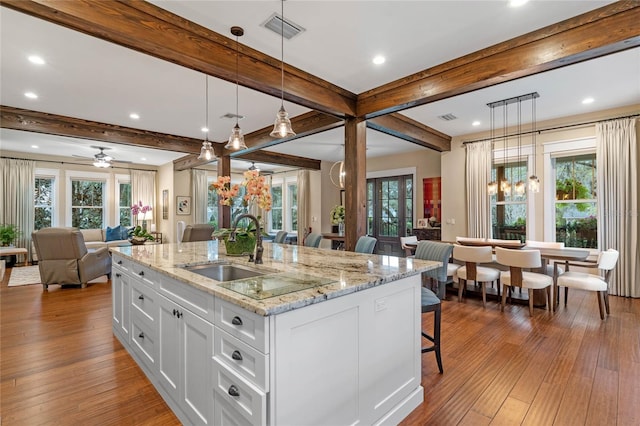 kitchen with light wood-type flooring, decorative light fixtures, beamed ceiling, white cabinetry, and an island with sink