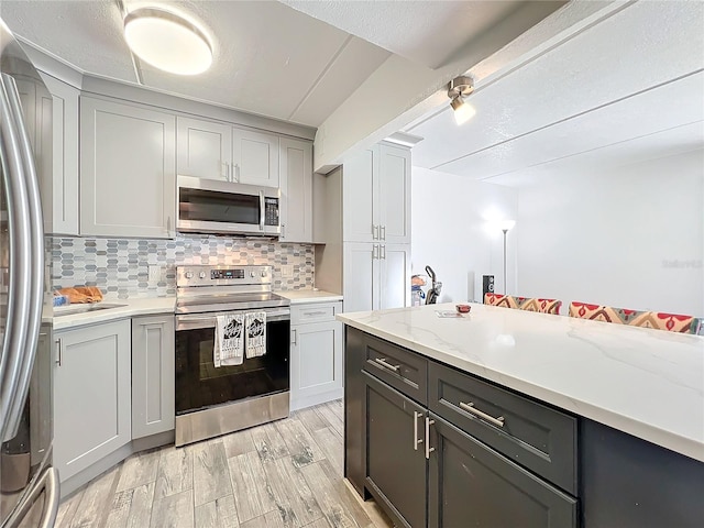 kitchen with gray cabinetry, backsplash, light wood-type flooring, appliances with stainless steel finishes, and light stone counters