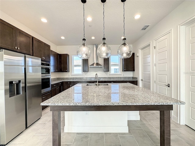 kitchen with wall chimney exhaust hood, a textured ceiling, stainless steel appliances, decorative light fixtures, and an island with sink