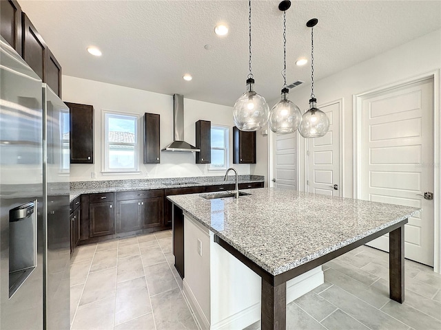 kitchen featuring sink, wall chimney exhaust hood, stainless steel fridge, a textured ceiling, and an island with sink