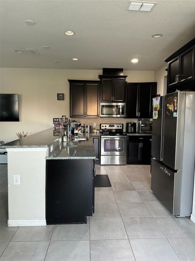 kitchen featuring sink, dark stone countertops, an island with sink, light tile patterned floors, and appliances with stainless steel finishes