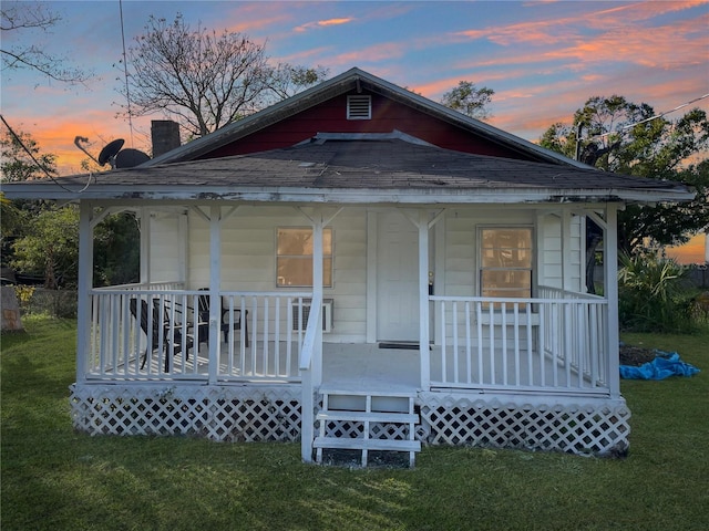 back house at dusk featuring covered porch and a lawn