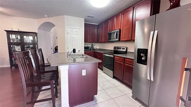 kitchen with sink, stainless steel appliances, an island with sink, a textured ceiling, and light tile patterned floors