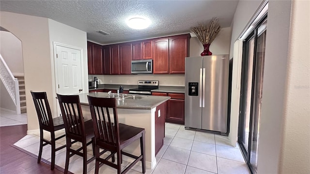 kitchen with a textured ceiling, stainless steel appliances, sink, light tile patterned floors, and a center island with sink