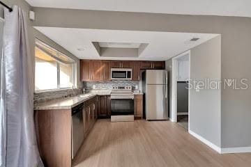 kitchen featuring decorative backsplash, stainless steel appliances, a tray ceiling, sink, and light hardwood / wood-style flooring