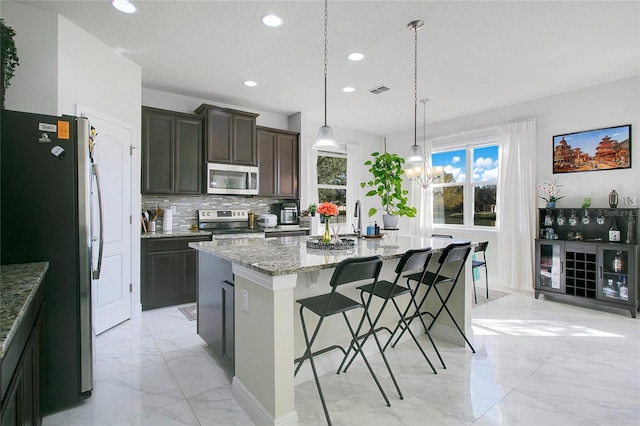 kitchen with light stone countertops, stainless steel appliances, an island with sink, decorative light fixtures, and dark brown cabinets