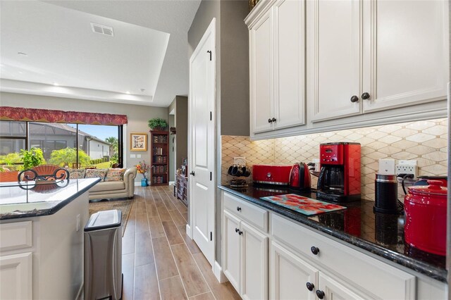 kitchen featuring light wood-type flooring, tasteful backsplash, a tray ceiling, dark stone countertops, and white cabinetry