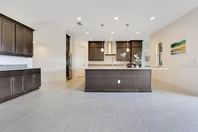 kitchen with dark brown cabinetry, wall chimney range hood, stainless steel refrigerator with ice dispenser, an island with sink, and pendant lighting