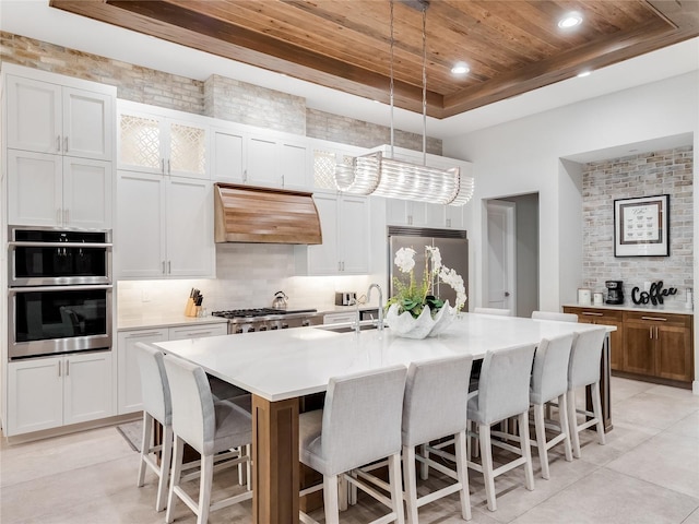 kitchen featuring appliances with stainless steel finishes, custom range hood, a center island with sink, decorative light fixtures, and white cabinets