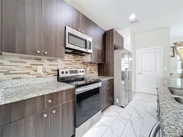 kitchen with light stone countertops, backsplash, a textured ceiling, stainless steel appliances, and dark brown cabinetry