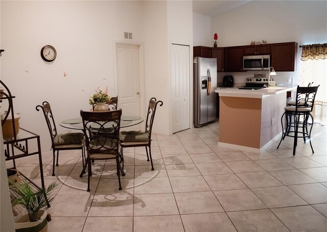 kitchen featuring high vaulted ceiling, appliances with stainless steel finishes, dark brown cabinets, light tile patterned flooring, and a breakfast bar area