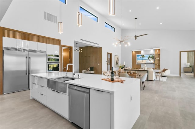 kitchen with ceiling fan with notable chandelier, white cabinets, appliances with stainless steel finishes, an island with sink, and hanging light fixtures