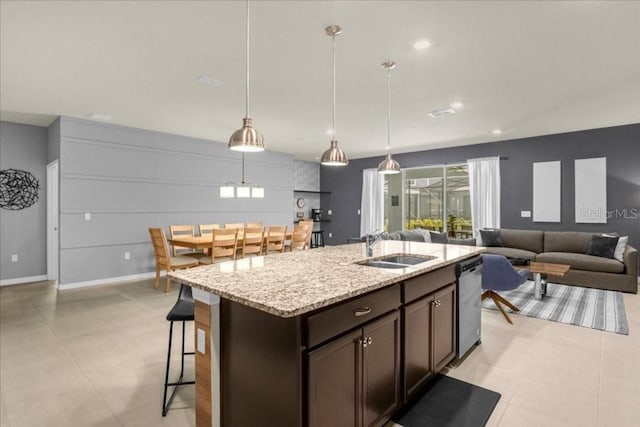 kitchen with a kitchen island with sink, sink, hanging light fixtures, dark brown cabinets, and light stone counters