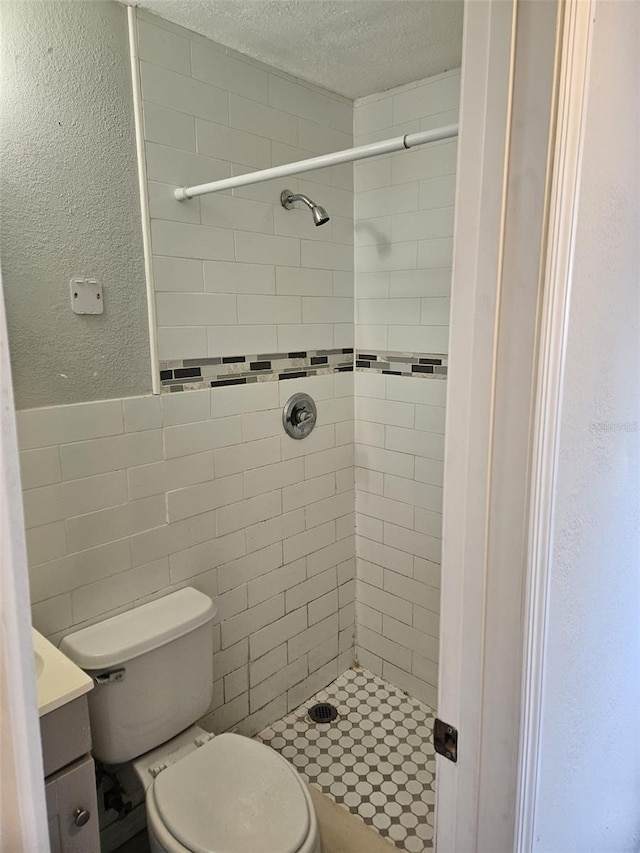 bathroom with a textured ceiling, vanity, tiled shower, tile walls, and toilet