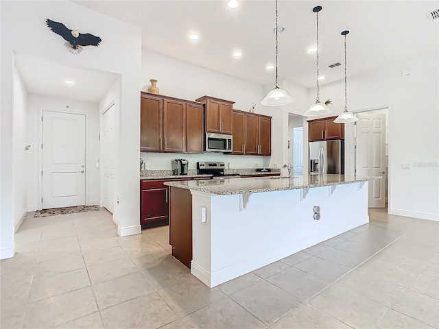 kitchen featuring a kitchen island with sink, light stone countertops, appliances with stainless steel finishes, decorative light fixtures, and a kitchen bar