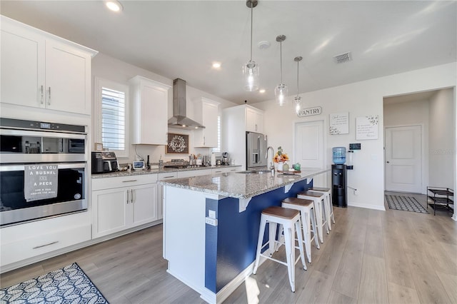 kitchen featuring white cabinets, wall chimney exhaust hood, an island with sink, and appliances with stainless steel finishes