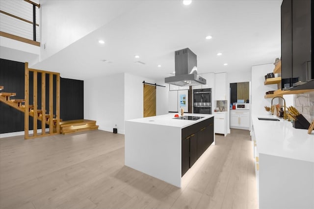 kitchen featuring an island with sink, white cabinetry, a barn door, island range hood, and double oven