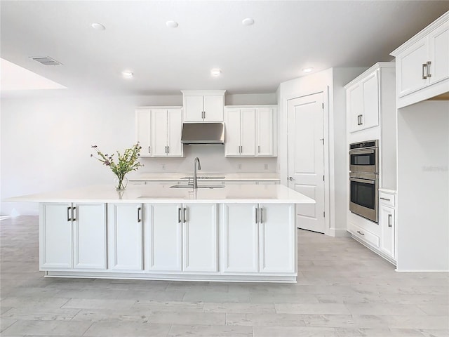 kitchen with double oven, white cabinetry, and a center island with sink