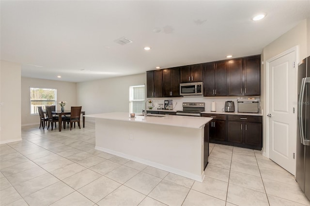 kitchen with dark brown cabinetry, stainless steel appliances, a kitchen island with sink, sink, and light tile patterned floors