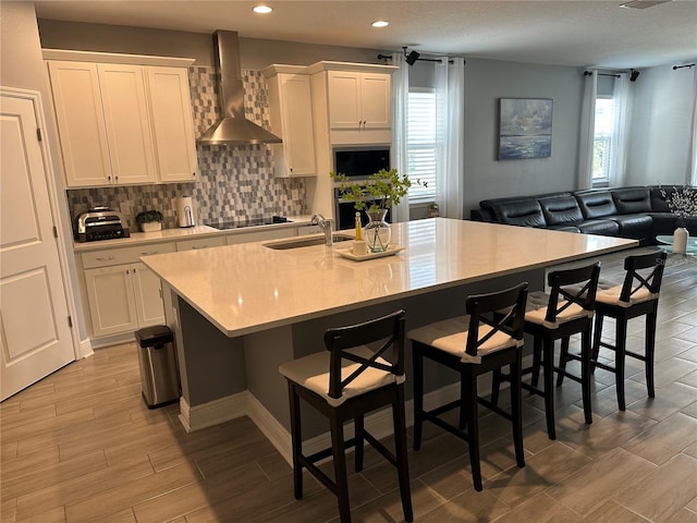 kitchen with wall chimney range hood, a sink, a kitchen island with sink, and white cabinets