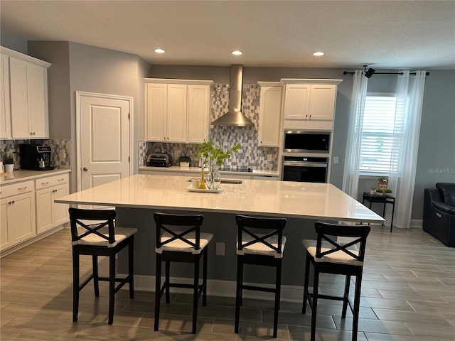 kitchen featuring light countertops, appliances with stainless steel finishes, white cabinets, wall chimney range hood, and an island with sink