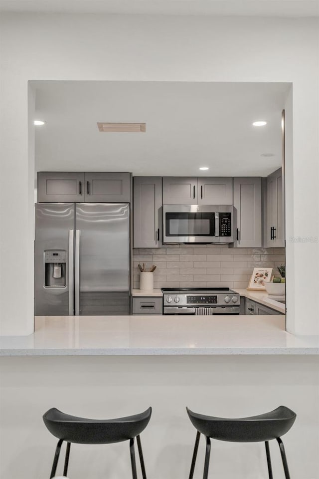 kitchen featuring light stone counters, gray cabinets, decorative backsplash, a breakfast bar, and appliances with stainless steel finishes