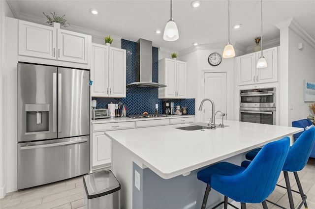 kitchen with appliances with stainless steel finishes, an island with sink, sink, white cabinets, and wall chimney range hood