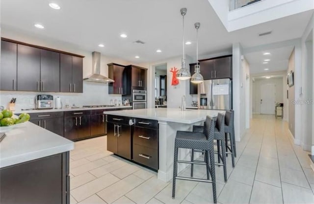 kitchen featuring wall chimney range hood, an island with sink, decorative light fixtures, dark brown cabinets, and appliances with stainless steel finishes