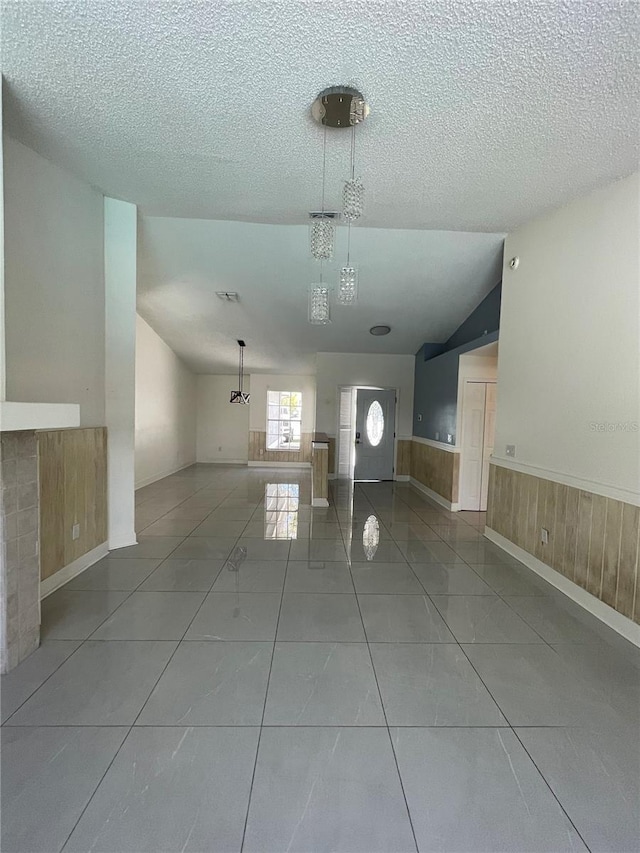 unfurnished living room featuring wooden walls, a wainscoted wall, vaulted ceiling, a textured ceiling, and tile patterned floors