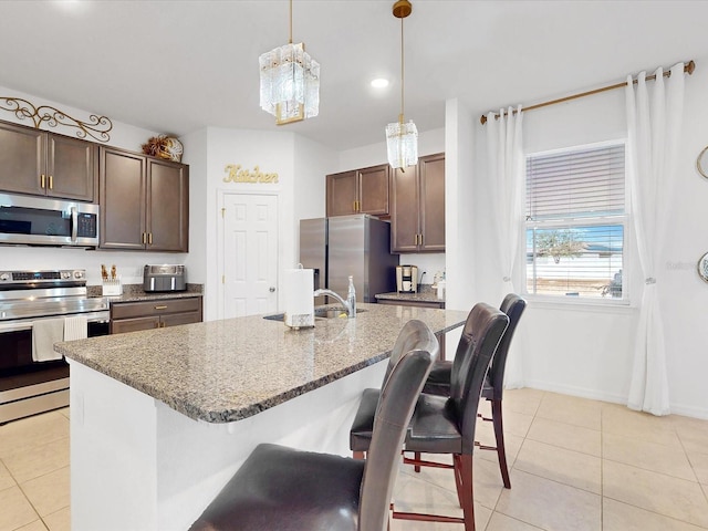 kitchen with light stone counters, dark brown cabinetry, stainless steel appliances, a kitchen island with sink, and decorative light fixtures