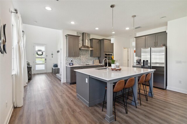 kitchen with wall chimney exhaust hood, stainless steel appliances, a kitchen island with sink, sink, and hanging light fixtures