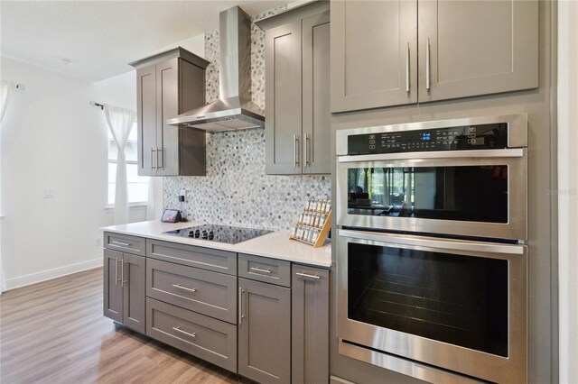 kitchen with black electric stovetop, wall chimney exhaust hood, gray cabinetry, stainless steel double oven, and light hardwood / wood-style flooring