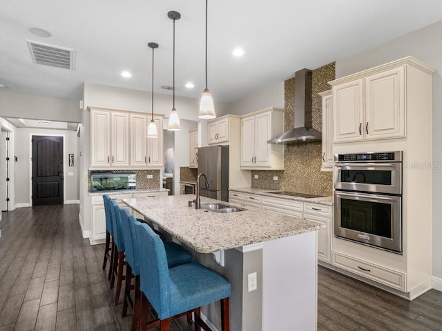 kitchen with pendant lighting, a kitchen island with sink, sink, wall chimney exhaust hood, and appliances with stainless steel finishes