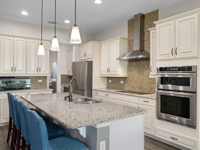 kitchen with a kitchen island with sink, sink, wall chimney exhaust hood, light stone counters, and stainless steel appliances