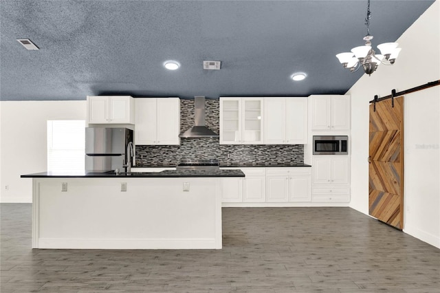 kitchen featuring appliances with stainless steel finishes, wall chimney range hood, a barn door, white cabinets, and hanging light fixtures