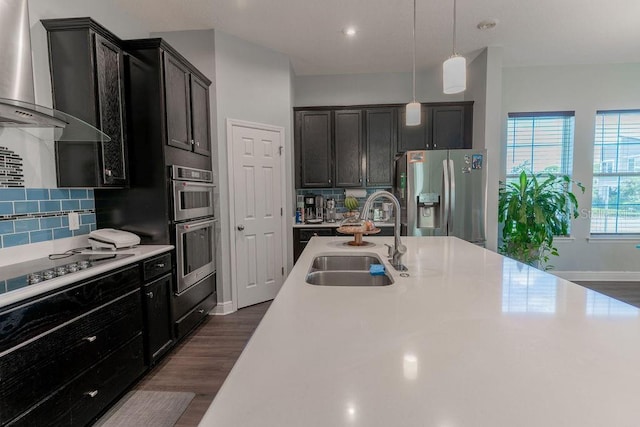 kitchen with pendant lighting, wall chimney exhaust hood, stainless steel appliances, sink, and backsplash