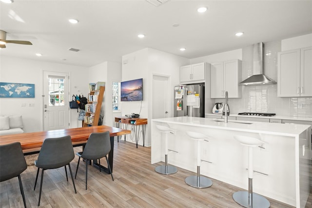 kitchen with stainless steel fridge, a center island with sink, white cabinetry, and wall chimney range hood