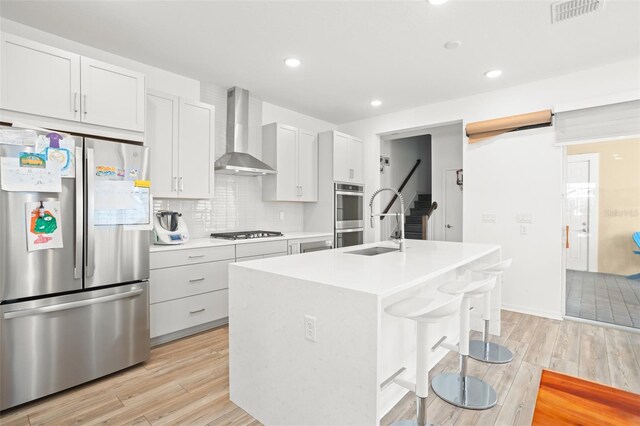 kitchen with white cabinetry, stainless steel appliances, wall chimney range hood, an island with sink, and light wood-type flooring