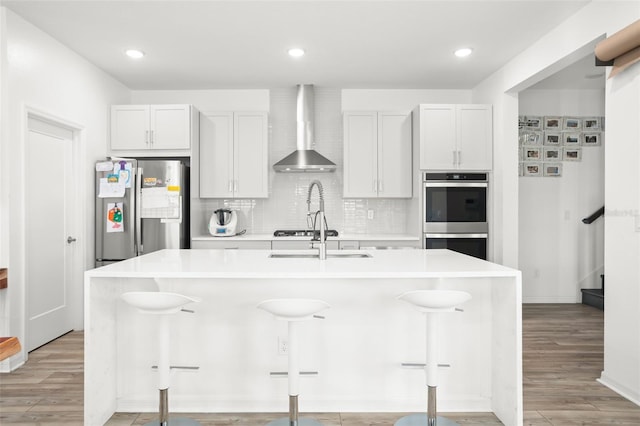 kitchen with white cabinets, wall chimney exhaust hood, stainless steel appliances, and an island with sink