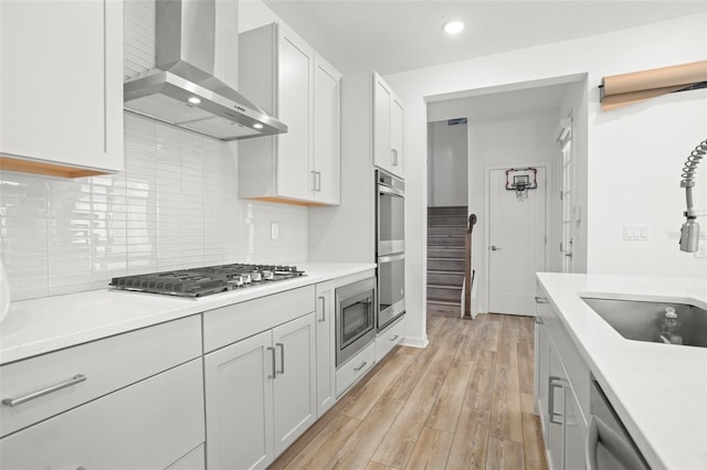 kitchen with white cabinets, wall chimney exhaust hood, light wood-type flooring, and appliances with stainless steel finishes