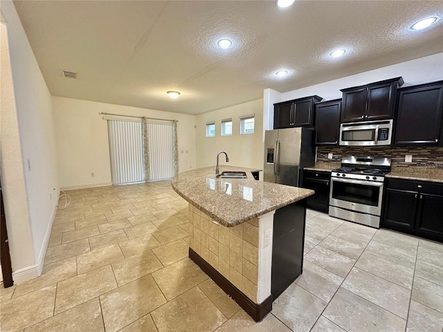 kitchen featuring stainless steel appliances, tasteful backsplash, sink, light stone counters, and a center island with sink