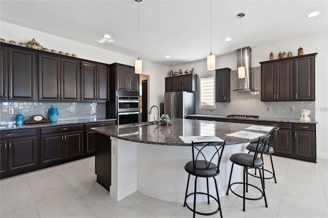 kitchen with a kitchen island with sink, pendant lighting, stainless steel appliances, and wall chimney range hood