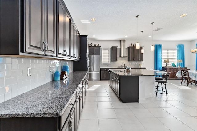 kitchen featuring stainless steel refrigerator, sink, wall chimney exhaust hood, an island with sink, and decorative light fixtures