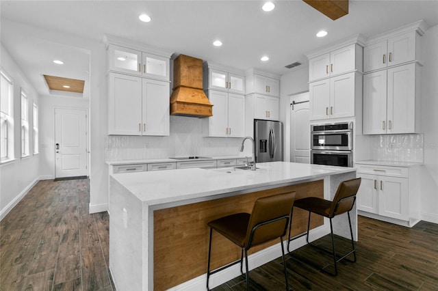 kitchen featuring appliances with stainless steel finishes, premium range hood, white cabinetry, and a center island with sink